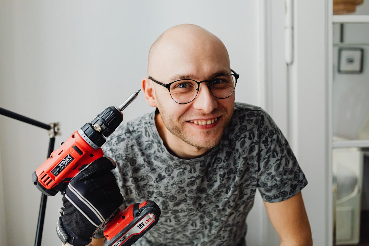 A smiling bald man with eyeglasses holding a red power drill indoors.